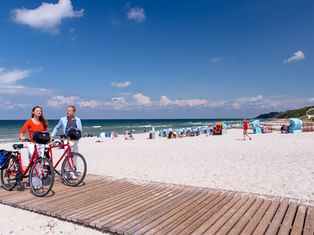 Zwei Radfahrer mit roten Rädern auf Holzsteg am Ostseestrand mit weißem Sand, blauen Strandkörben und Menschen an einem sonnigen Tag.