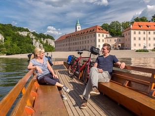 Zwei Radfahrer sitzen mit ihren Rädern auf einer Holzfähre und überqueren einen Fluss. Historische Gebäude mit roten Dächern und Kirchturm am Ufer.