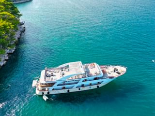 Luftaufnahme der MS Olmissum, ein weißes Kreuzfahrtschiff mit blauem Streifen, vor Anker in türkisem Wasser nahe bewaldeter Küste.