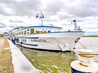 Weißes Flusskreuzfahrtschiff MS Rigoletto mit blauem Rand am Betonpier vertäut. Das Schiff hat große Fenster und Flaggen auf dem Oberdeck.