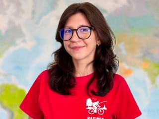 Krystsina mit dunklen, welligen Haaren und Brille trägt ein rotes Radweg-Reisen T-Shirt und lächelt vor einer bunten Weltkarte.