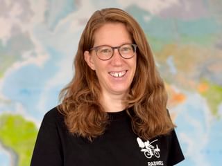 Damaris mit Brille und langen braunen Haaren, lächelnd vor einer Weltkarte. Sie trägt ein schwarzes Shirt mit Fahrrad-Logo.