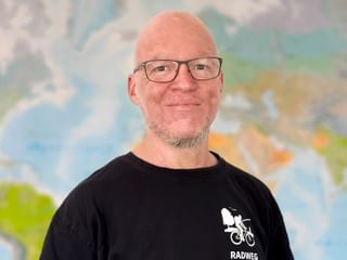 Portrait von Stefan G. mit Brille und schwarzem Radweg-Shirt, lächelnd vor einem bunten Weltkarten-Hintergrund.