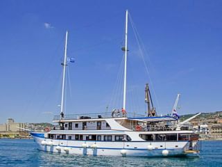 Weiße Motoryacht MS Aria mit blauem Zierstreifen und zwei Masten im Hafen. Weiße Fender entlang des Rumpfes, Küstenstadt im Hintergrund.
