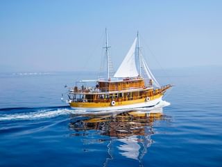 MS Planka, ein hölzernes Segelschiff mit weißen Segeln und zwei Masten, fährt auf ruhigem blauen Wasser mit fernen Bergen am Horizont.