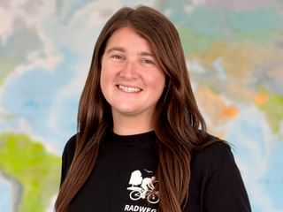 Porträt von Michelle mit langen braunen Haaren, die warm lächelt. Sie trägt ein schwarzes Shirt mit Fahrrad-Logo. Im Hintergrund ist eine Weltkarte sichtbar.