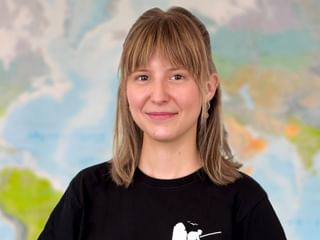 Porträt von Celine mit schulterlangen blonden Haaren und Pony, in schwarzem Shirt, lächelnd vor einer Weltkarte im Hintergrund.