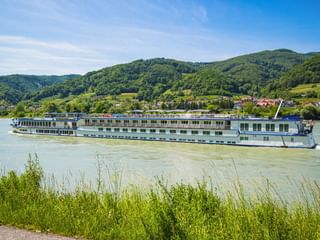 Weißes Flusskreuzfahrtschiff MS Swiss Crown auf der Donau mit grünen Hügeln, einem Dorf und blauem Himmel im Hintergrund.