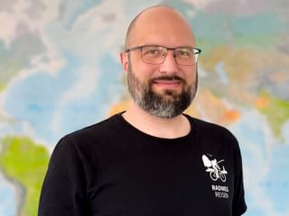 Porträt von Max mit Brille und schwarzem Radweg Reisen Shirt mit Fahrrad-Logo, stehend vor einer bunten Weltkarte.