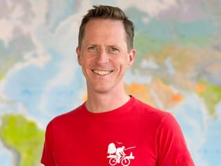 Thomas trägt ein rotes Radtrikot mit Fahrrad-Logo und lächelt in die Kamera. Weltkarte im unscharfen Hintergrund sichtbar.