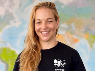 Anja mit langen blonden Haaren lächelnd, trägt ein schwarzes Radweg-Shirt mit Radfahrer-Logo. Im Hintergrund ist eine bunte Weltkarte sichtbar.