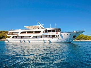 Weiße Motoryacht MY Mystic ankert in ruhigem blauen Wasser mit grüner Küstenlinie im Hintergrund unter klarem Himmel.