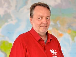Porträt von Claus in einem roten Poloshirt mit Fahrrad-Logo, stehend vor einer bunten Weltkarte im Hintergrund.