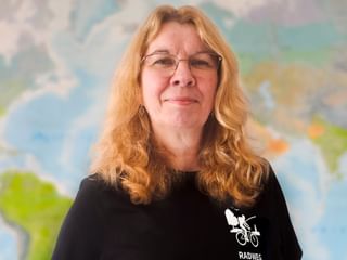 Porträt von Adriana mit Brille und welligem blondem Haar, trägt ein schwarzes Shirt mit Fahrrad-Logo, steht vor einer Weltkarte.