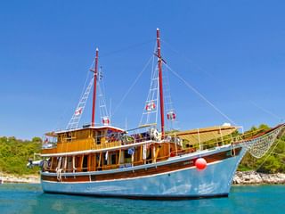 MS Orkan, ein Holzkreuzfahrtschiff mit blauem Rumpf und zwei roten Masten, vor Anker in türkisfarbenem Wasser nahe bewaldeter Küste unter blauem Himmel.