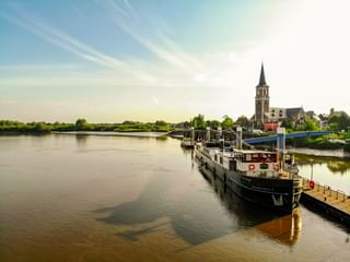 Flusskreuzfahrtschiff MS Magnifique II an einem Holzsteg vertäut. Eine Kirche mit hohem Turm steht am gegenüberliegenden Flussufer unter blauem Himmel.
