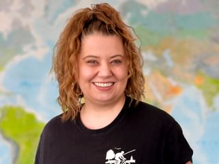 Yana mit lockigem braunem Haar und gelben Ohrringen, breit lächelnd in einem schwarzen Fahrrad-T-Shirt. Im Hintergrund ist eine bunte Weltkarte sichtbar.