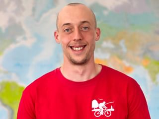 Porträt von Tim S in einem roten Shirt mit Fahrrad-Logo, lächelnd in die Kamera. Im Hintergrund ist eine bunte Karte sichtbar.