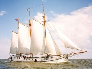 Dreimastklipper Elizabeth segelt mit weißen Segeln vor blauem Himmel mit Wolken. Das weiße Schiff bewegt sich durch ruhiges Wasser.