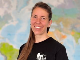 Frau mit langen braunen Haaren in schwarzem Rad-T-Shirt, lächelt in die Kamera. Bunte Weltkarte im Hintergrund sichtbar.