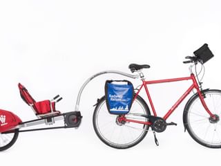 Rotes Tourenrad mit blauen Packtaschen und Lenkertasche, verbunden mit einem roten WeeHoo Einrad-Kinderanhänger mit Sitz und Sicherheitswimpel.