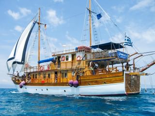 MS Panagiota, ein hölzernes Segelschiff mit weißem Rumpf und zwei Masten, segelt auf blauem Meer unter klarem Himmel mit griechischer Flagge.