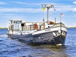 MS Flora, ein weiß-schwarzes Fahrgastschiff mit blauen Fenstern, fährt auf ruhigem Wasser unter blauem Himmel mit weißen Wolken.