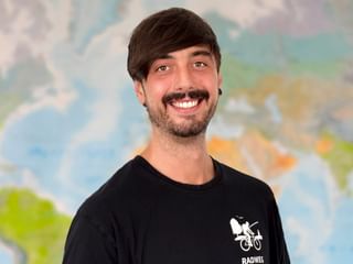 Porträt von Marian, Teammitglied von Radweg-Reisen, mit dunklen Haaren und Schnurrbart, lächelnd vor einer Weltkarte. Er trägt ein schwarzes Shirt.