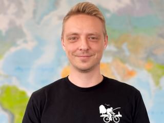 Portrait von Raul in schwarzem T-Shirt mit Fahrrad-Logo, lächelnd vor einer Weltkarte im Hintergrund.