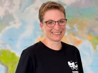 Beate mit Brille und schwarzem Shirt mit Fahrrad-Logo, lächelnd vor einer bunten Weltkarte.
