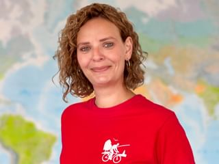 Ilka mit lockigem braunen Haar trägt ein rotes Shirt mit Fahrrad-Logo und lächelt vor einem unscharfen Weltkartenhintergrund.