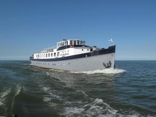 Weiß-dunkelblaues Passagierschiff Floor van Aemstel fährt auf offenem Wasser unter klarem blauen Himmel und erzeugt eine Bugwelle.