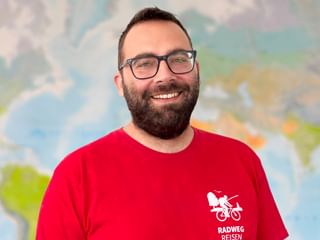 Portrait von Tim J. mit Brille und rotem Radweg Reisen Shirt, lächelnd vor einem Weltkarten-Hintergrund.