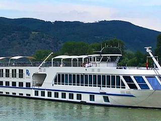 Weißes Flusskreuzfahrtschiff MS SE-Manon mit blauem Streifendesign fährt auf ruhigem Wasser vor grünen Bergen.