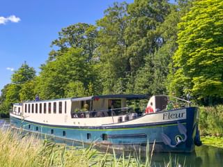 Flusskreuzfahrtschiff MS Fleur an einem von Bäumen gesäumten Kanal in Frankreich. Das weiß-blaue Schiff hat ein offenes Deck mit Passagieren.