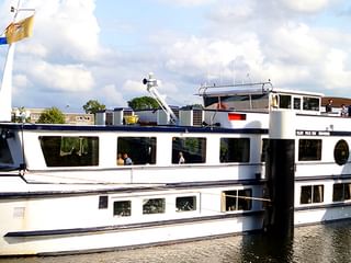 Weißes Flusskreuzfahrtschiff MS Olympia am Kai mit wehenden Flaggen. Weitere Schiffe im Hintergrund unter bewölktem Himmel.