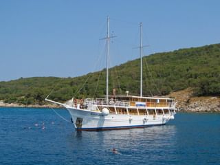 Weißes Motorsegelschiff MS Kazimir mit zwei Masten vor Anker in ruhigem blauen Wasser. Grüne Hügel im Hintergrund, Schwimmer im Wasser.