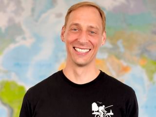 Porträt von Andreas in einem schwarzen T-Shirt mit Radfahrer-Grafik, der breit lächelt vor einem unscharfen Weltkarten-Hintergrund.