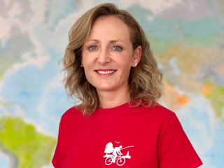 Portrait von Verena in rotem Shirt mit Radfahrer-Logo, lächelnd vor einer Weltkarte im Hintergrund.