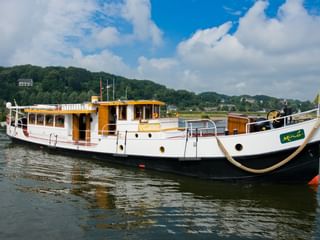 MS Miro, ein weiß-gelbes Flusskreuzfahrtschiff mit schwarzem Rumpf, liegt vor Anker auf ruhigem Wasser mit grünen Hügeln im Hintergrund.