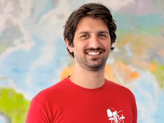 Arthur mit dunklen Haaren und Bart, trägt ein rotes T-Shirt mit Radfahrer-Logo, lächelt vor einer bunten Weltkarte.