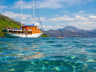 MS Mirabela, ein hölzernes Segelschiff mit weißen Masten, ankert in kristallklarem türkisfarbenem Wasser. Berge und grüne Hügel im Hintergrund.
