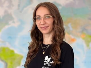 Porträt von Nina mit Brille und langen welligen braunen Haaren, trägt ein schwarzes Shirt mit Radfahrer-Grafik. Eine Weltkarte ist im Hintergrund sichtbar.