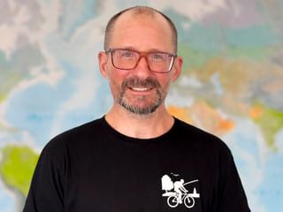 Porträt von Lars mit Brille und schwarzem Shirt mit Fahrrad-Logo, lächelnd vor einer Weltkarte im Hintergrund.