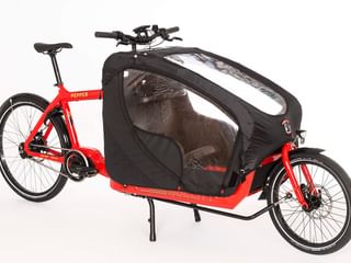 Rotes Lastenrad mit geschlossener schwarzer Stoffkabine mit transparenten Fenstern. Das Rad hat zwei Räder vorne und eins hinten mit Ständer.