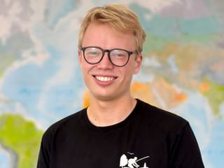 Peter mit blonden Haaren und schwarzer Brille, der breit lächelt. Er trägt ein schwarzes T-Shirt. Im Hintergrund ist eine bunte Karte sichtbar.