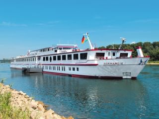 Weißes Flusskreuzfahrtschiff MS Normandie mit rotem Rand fährt auf blauem Wasser unter klarem Himmel, grüne Bäume und felsiges Ufer sichtbar.