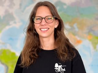 Annika mit Brille und langen braunen Haaren, lächelnd vor einer Weltkarte. Sie trägt ein schwarzes Shirt mit Fahrrad-Logo.