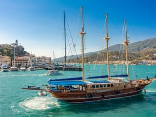 Holzsegelschiff MS Thalassa mit vier Masten in türkisfarbenem Hafenwasser, mit weißer Hangstadt und Bergen im Hintergrund.