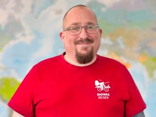 Porträt von Alex mit Brille und rotem Radweg Reisen T-Shirt mit Firmenlogo, lächelnd vor einer Weltkarte im Hintergrund.
